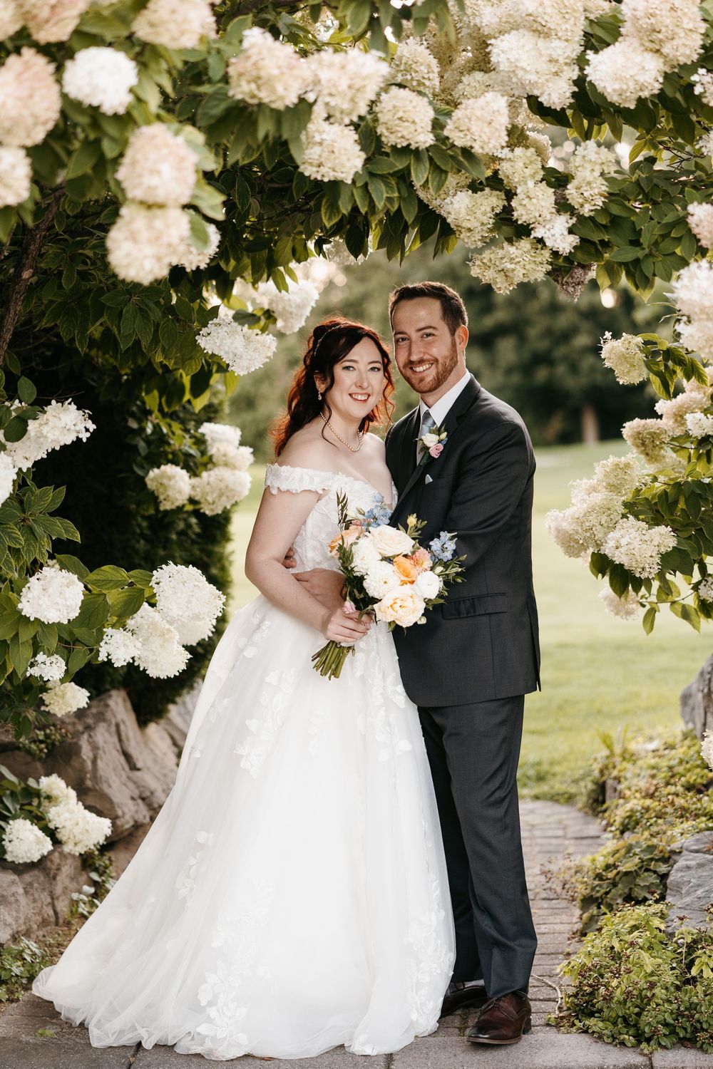 Elegant wedding couple poses under blooming white floral arbor in garden setting with flowing bridal gown.