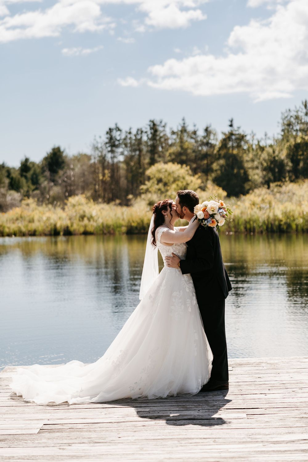 A couple embraces on a wooden dock beside a serene lake with trees in the background on their wedding day.