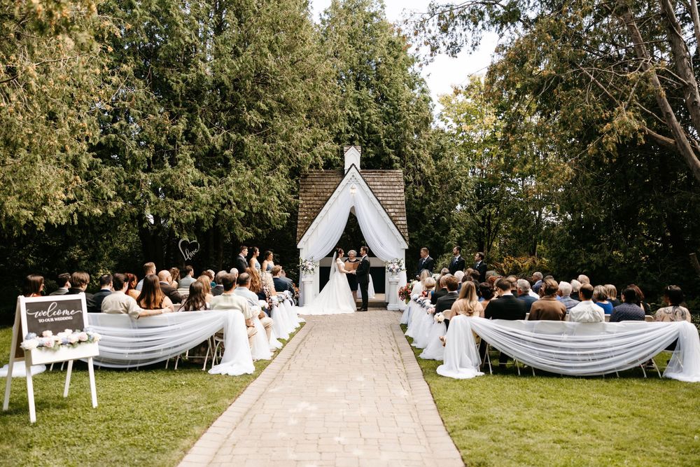 A panoramic view of a scenic outdoor wedding ceremony with white decorations set in a lush green garden setting.