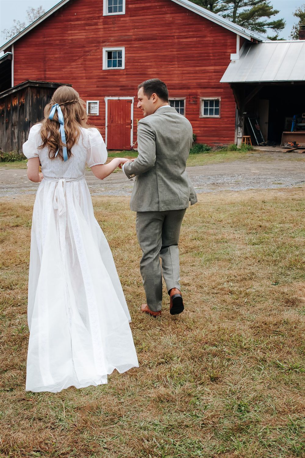 Two people in wedding attire walk playfully in front of a rustic red barn.