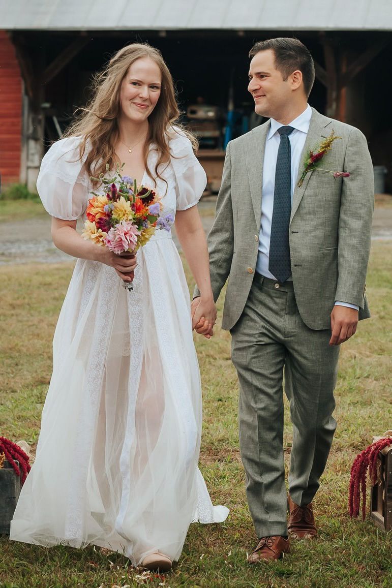 Couple in white dress and grey suit walking together outdoors holding autumn-colored bouquets.