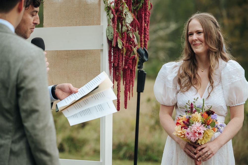 A wedding couple embraces at a rustic outdoor ceremony under hanging burgundy streamers and a white arbor.