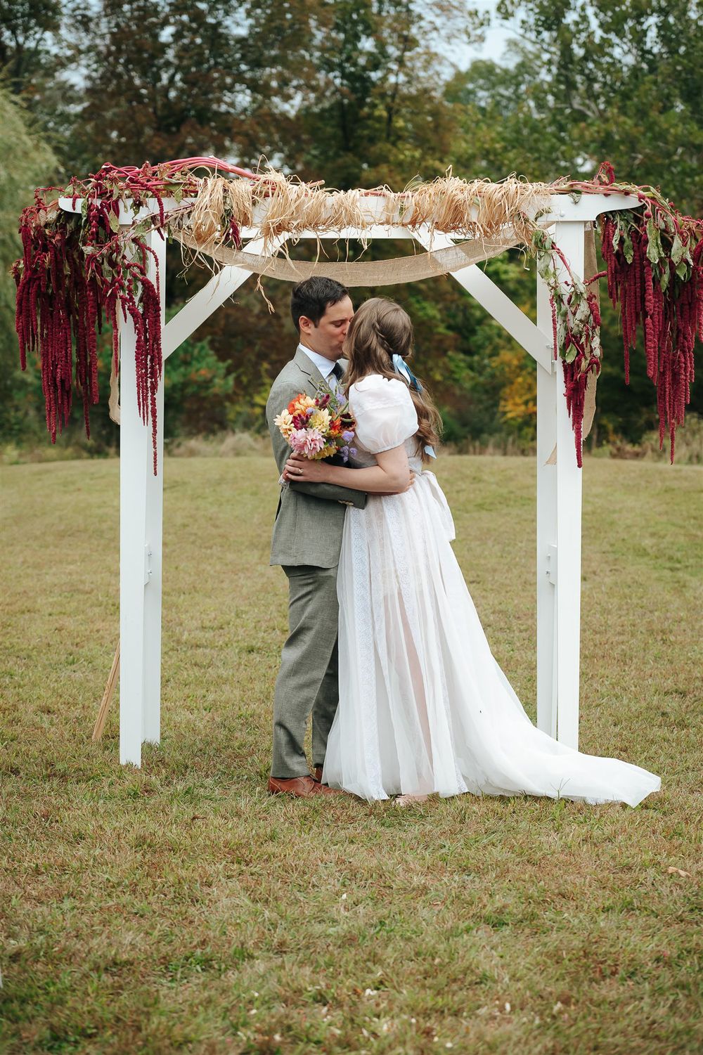 An outdoor wedding ceremony setup features a white wooden arbor decorated with flowing burgundy ribbons on a grassy hill.