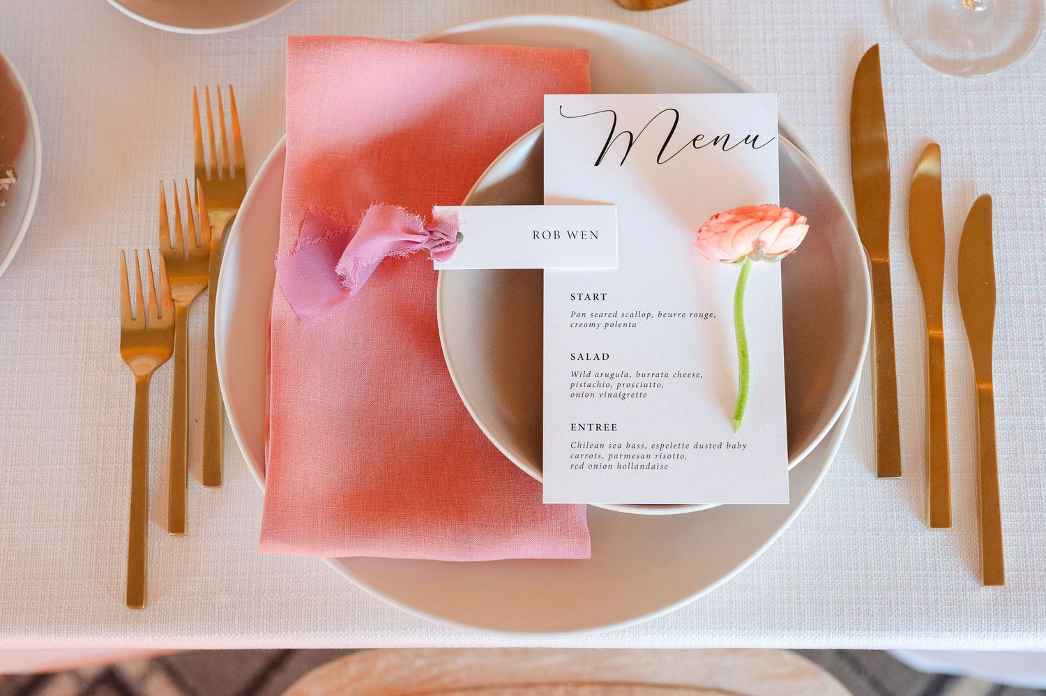 An elegant place setting with gold cutlery, pink napkin, and a menu card decorated with fresh coral flowers.