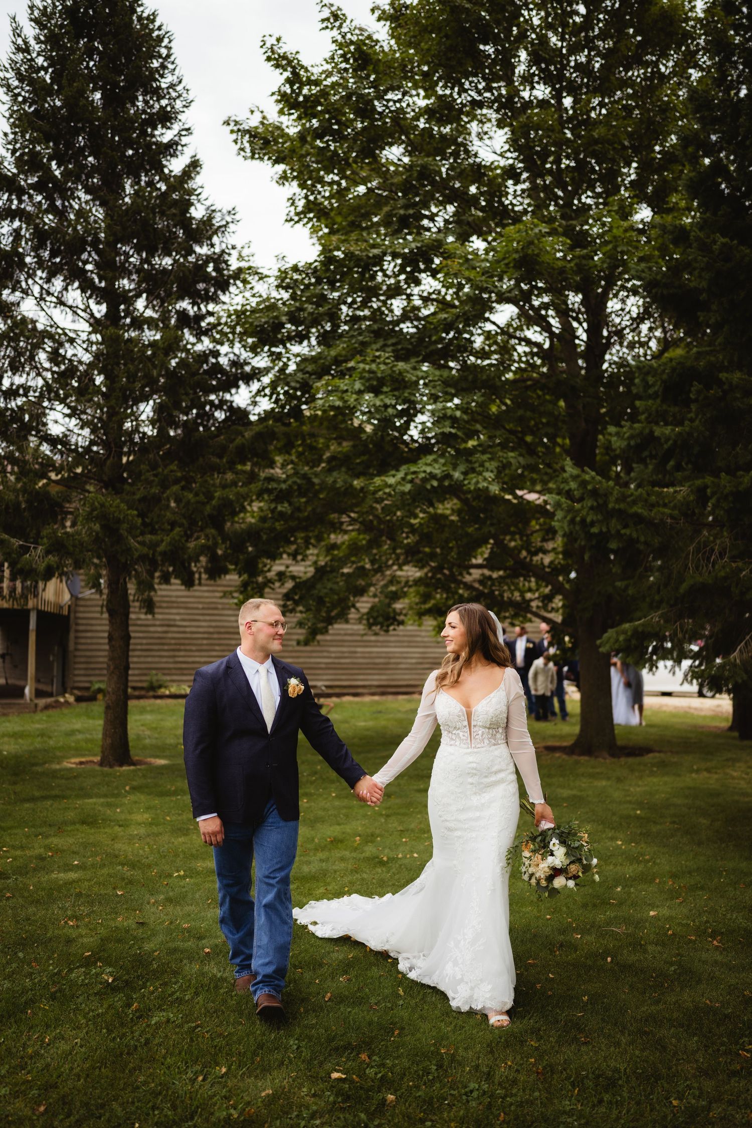 A couple in wedding attire dances together on a grassy lawn surrounded by trees during an outdoor celebration.