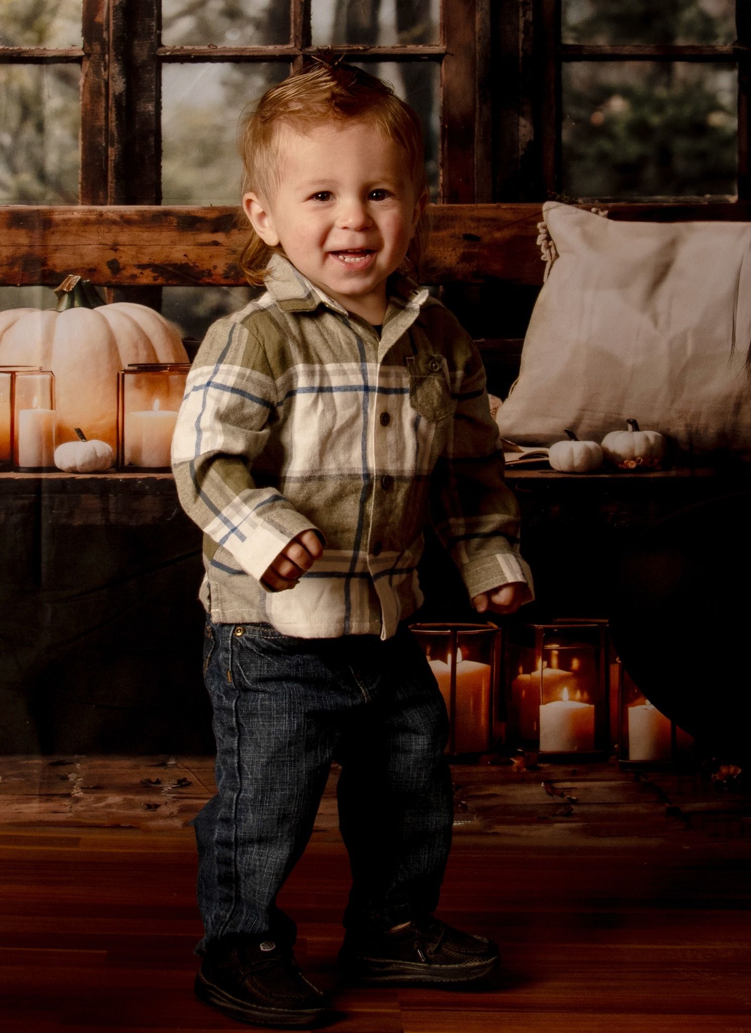 Sweet toddler in a plaid sweater and jeans sitting and playing on a rustic wooden floor with autumn decor.