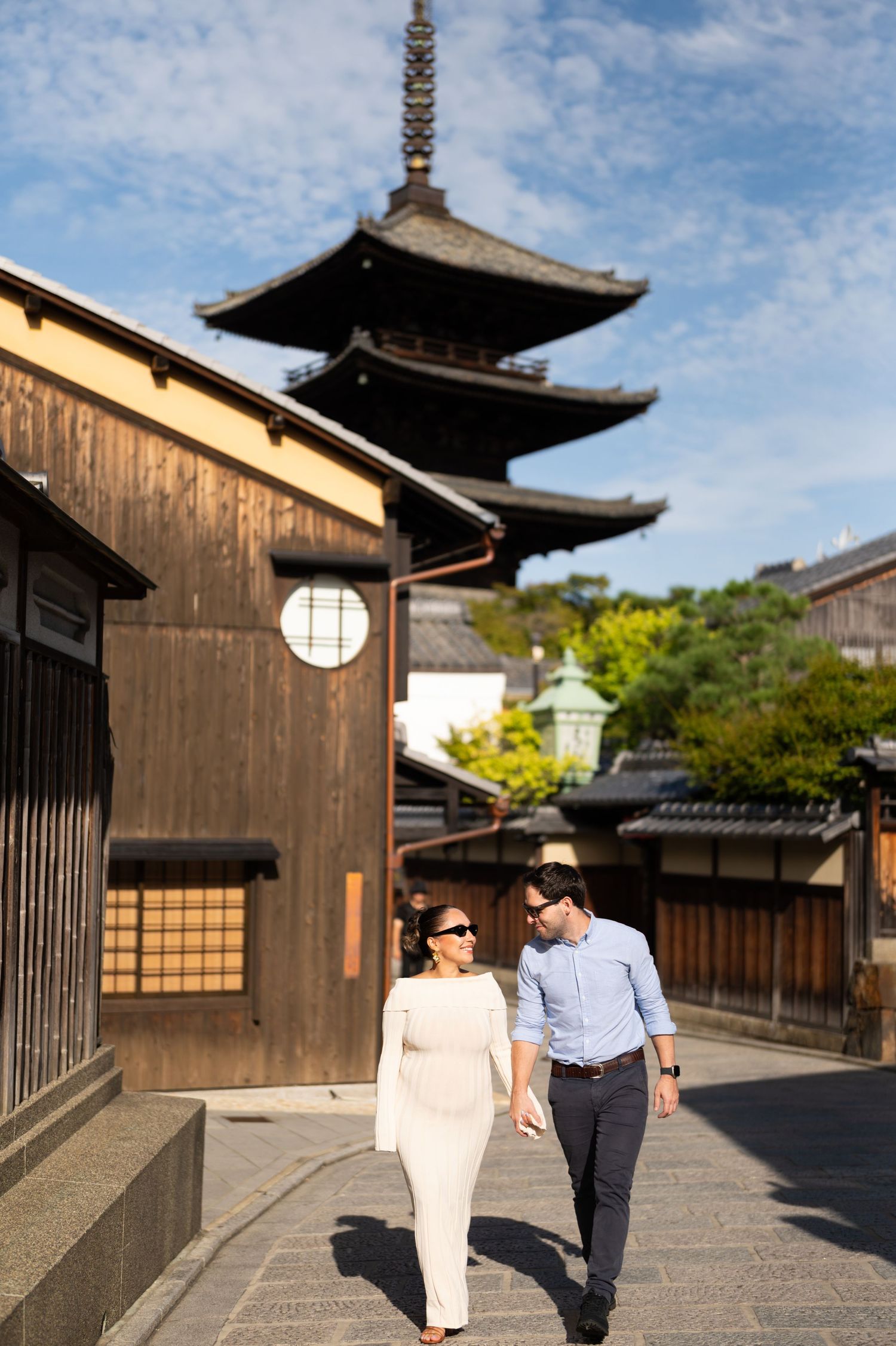 Pregnant couple walks hand-in-hand on a cobblestone street with a Japanese pagoda backdrop.