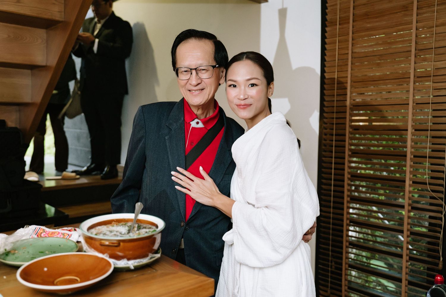 Elderly man in red-accented jacket embracing bride in white robe near food spread indoors.
