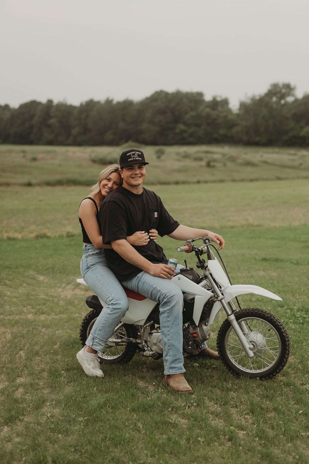 A couple sits together on a white dirt bike motorcycle in a grassy field on an overcast day.