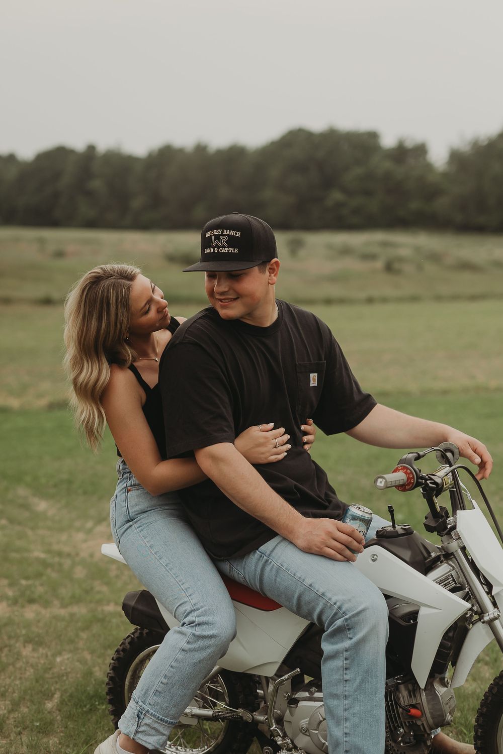 A couple shares an intimate moment while sitting on a motorcycle in a rural field setting.