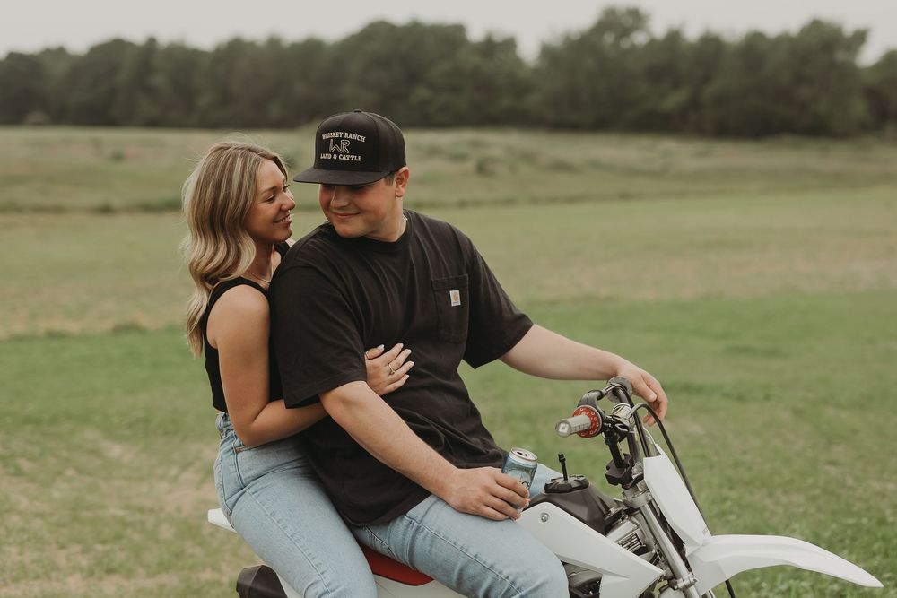 Two people embrace while sitting on a motorcycle in a green field during sunset.