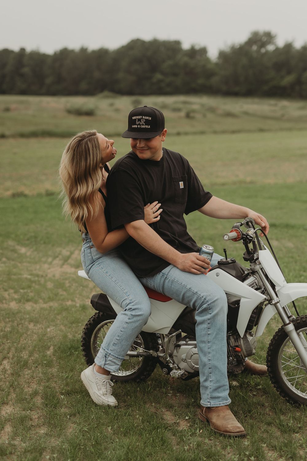 A couple sits together on a white dirt bike in a grassy field during a romantic outdoor photo session.