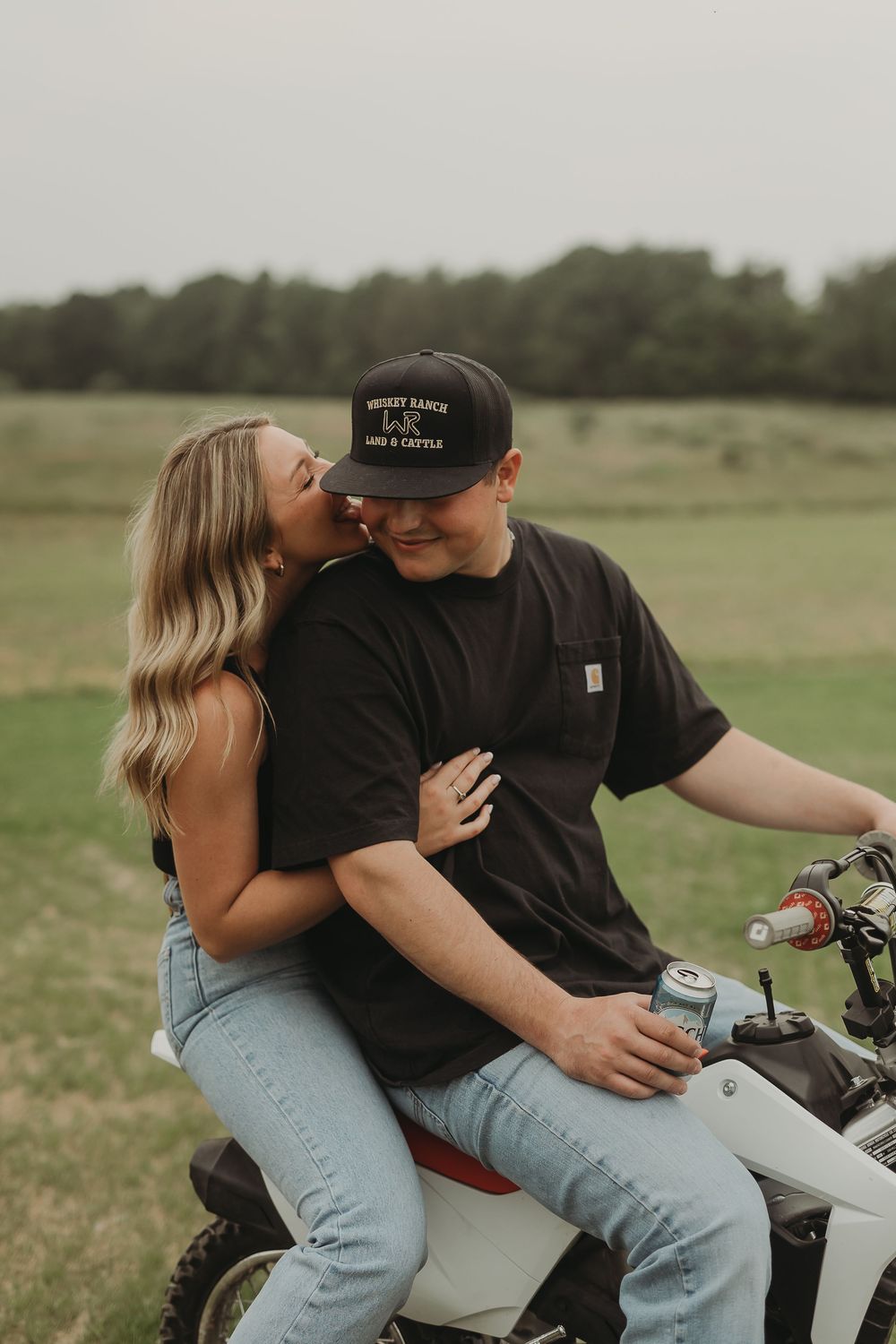 A couple shares an intimate moment on a dirt bike in an open field with trees in the background.