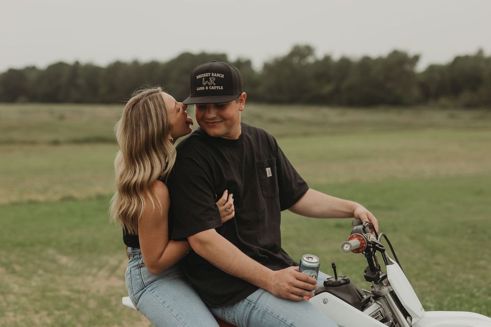A couple embraces next to a vintage motorcycle in a serene green field during sunset.