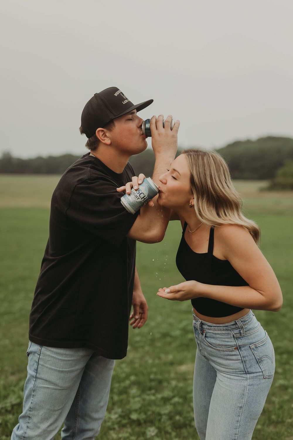 A playful couple drinks from water bottles together in a grassy field on an overcast day.