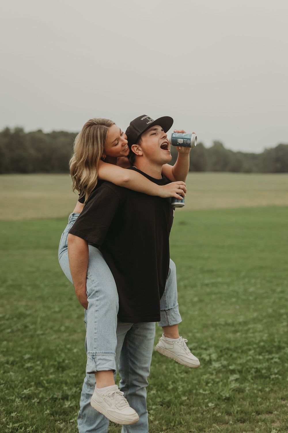 Playful couple embraces in a grassy green field on an overcast day, showing joyful affection and laughter.