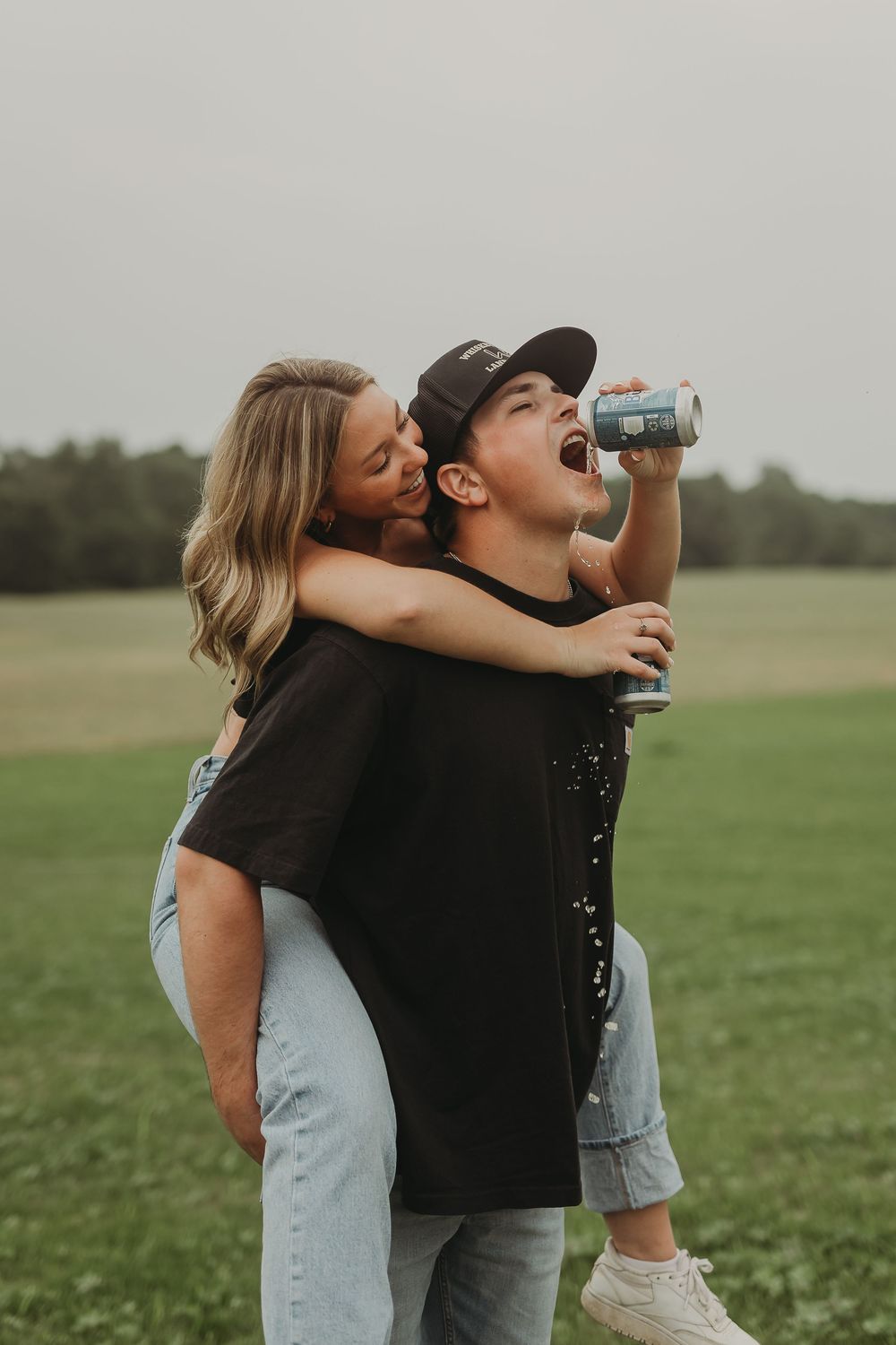 A playful couple having fun in a grassy field with one getting a piggyback ride while the other drinks from a can.
