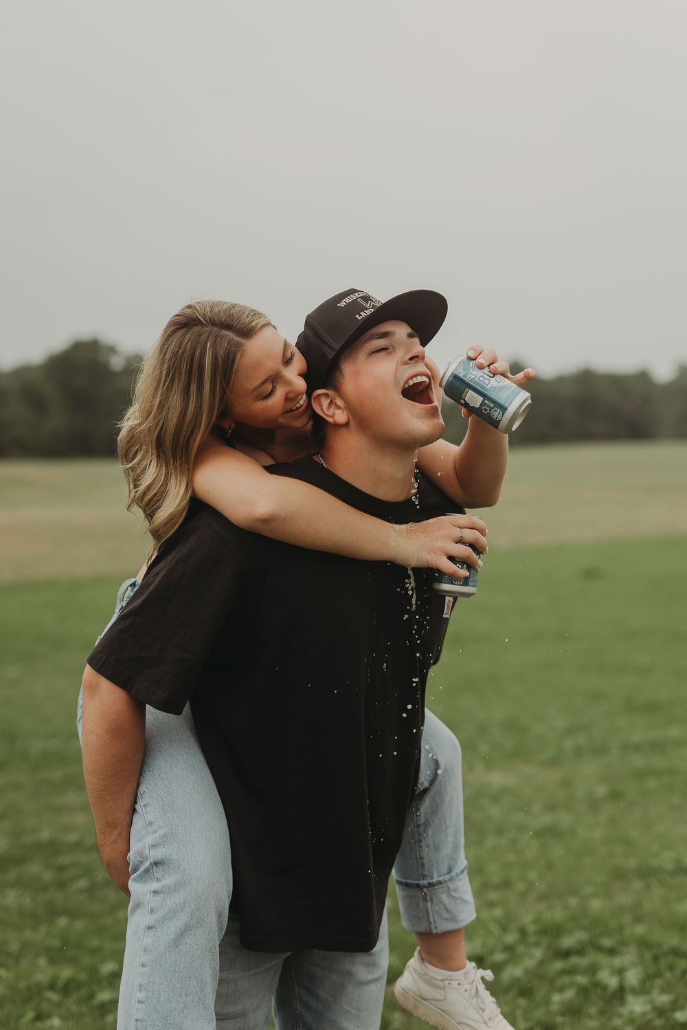 A playful couple shares a drink from a metal bottle while enjoying time together in a grassy field.