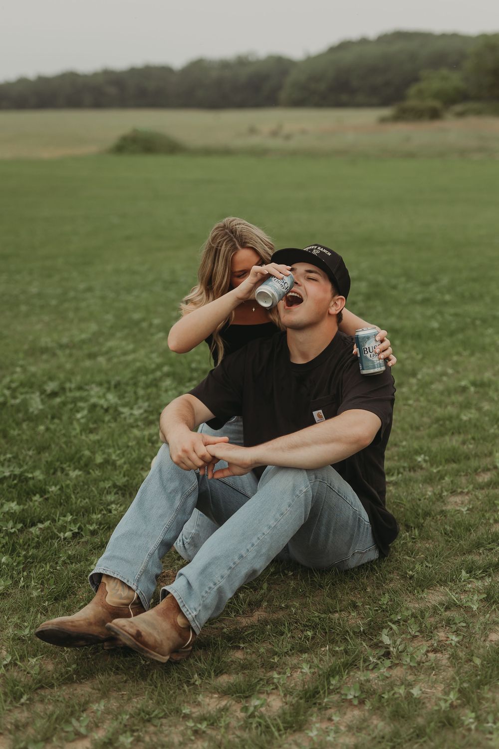 A couple in matching black shirts and jeans sharing a drink together while sitting in a grassy field.