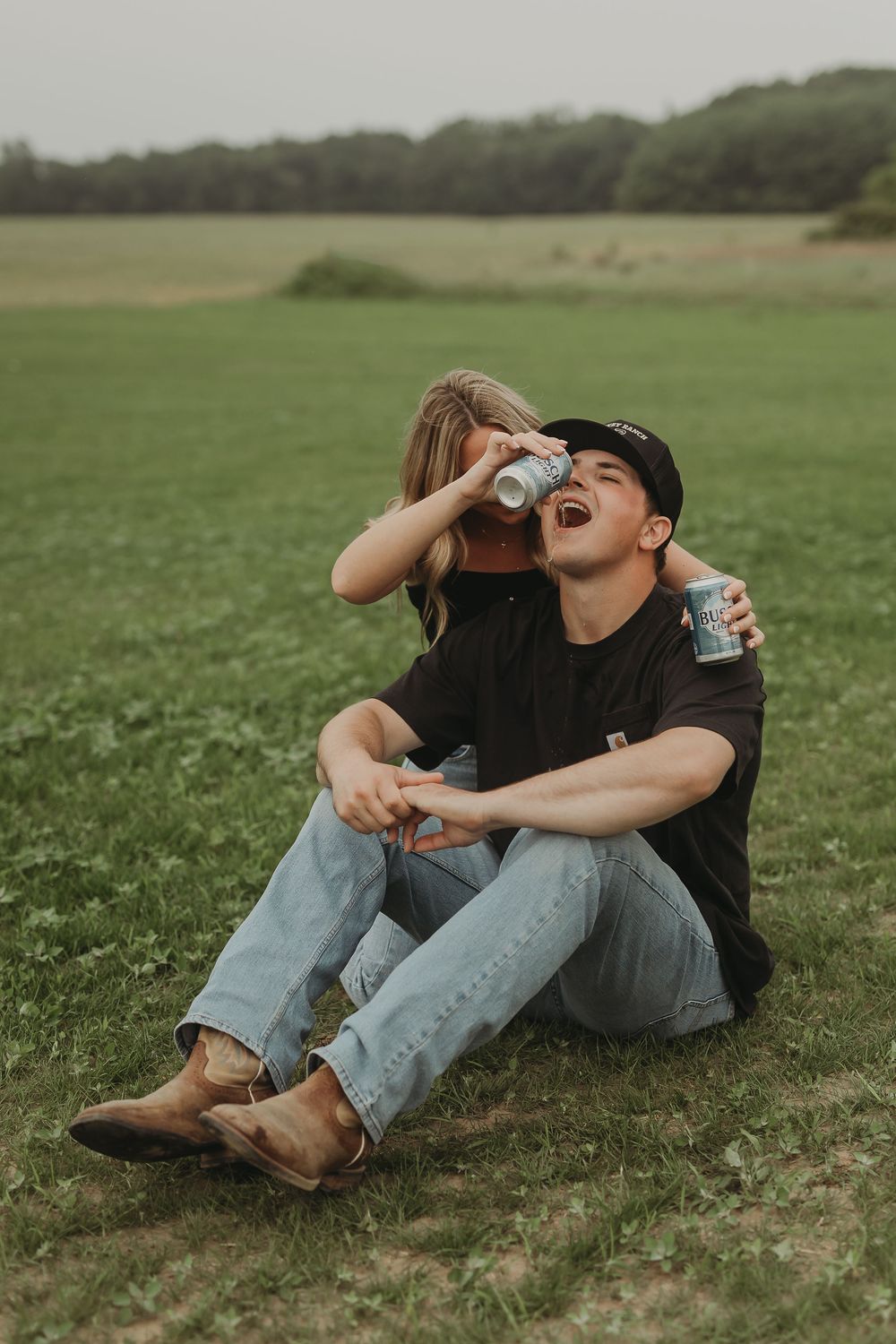 A couple shares intimate laughter while sitting close together on grassy meadow in coordinated casual outfits.
