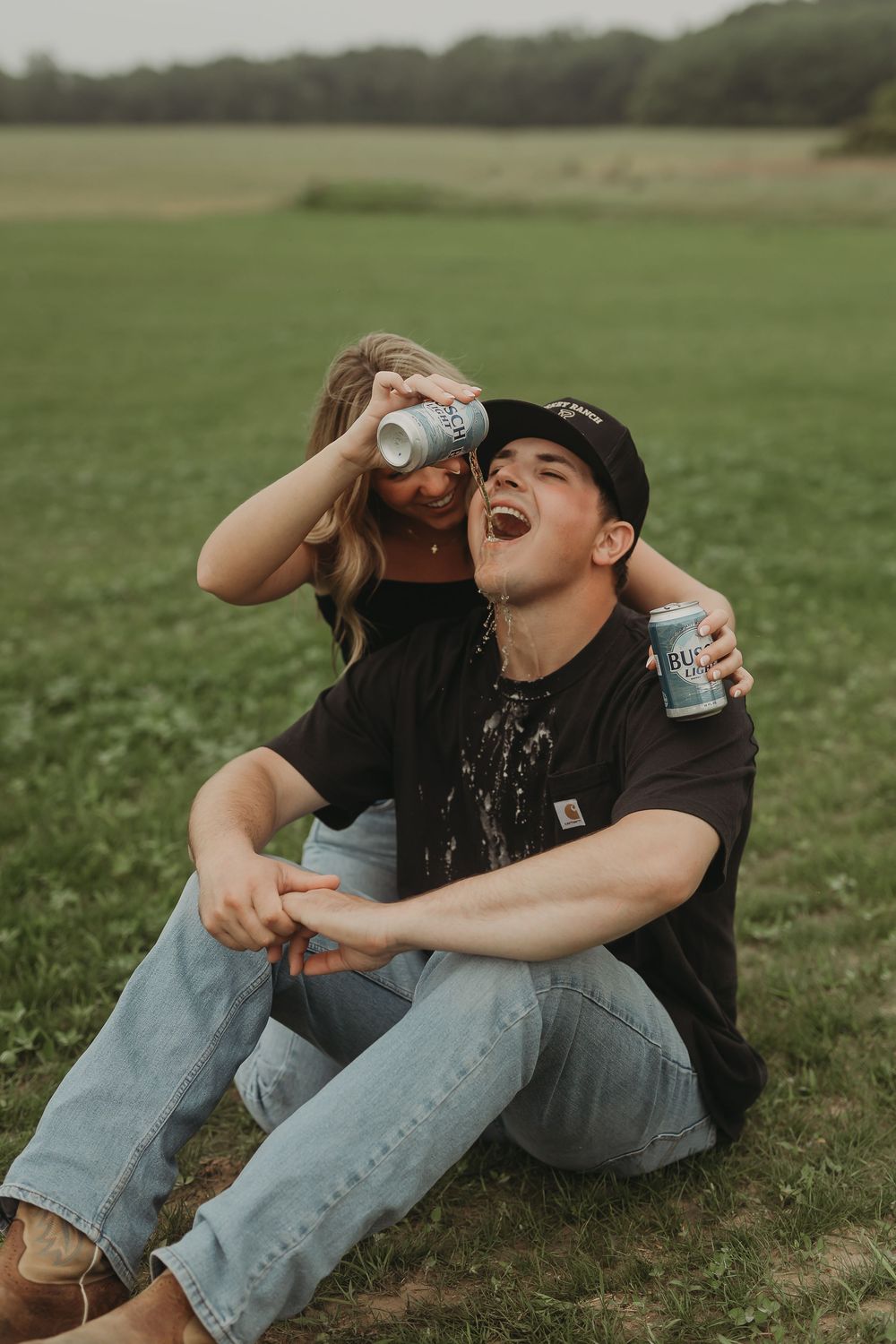 A couple sits together on grass while laughing and drinking from glass bottles on a serene summer day.