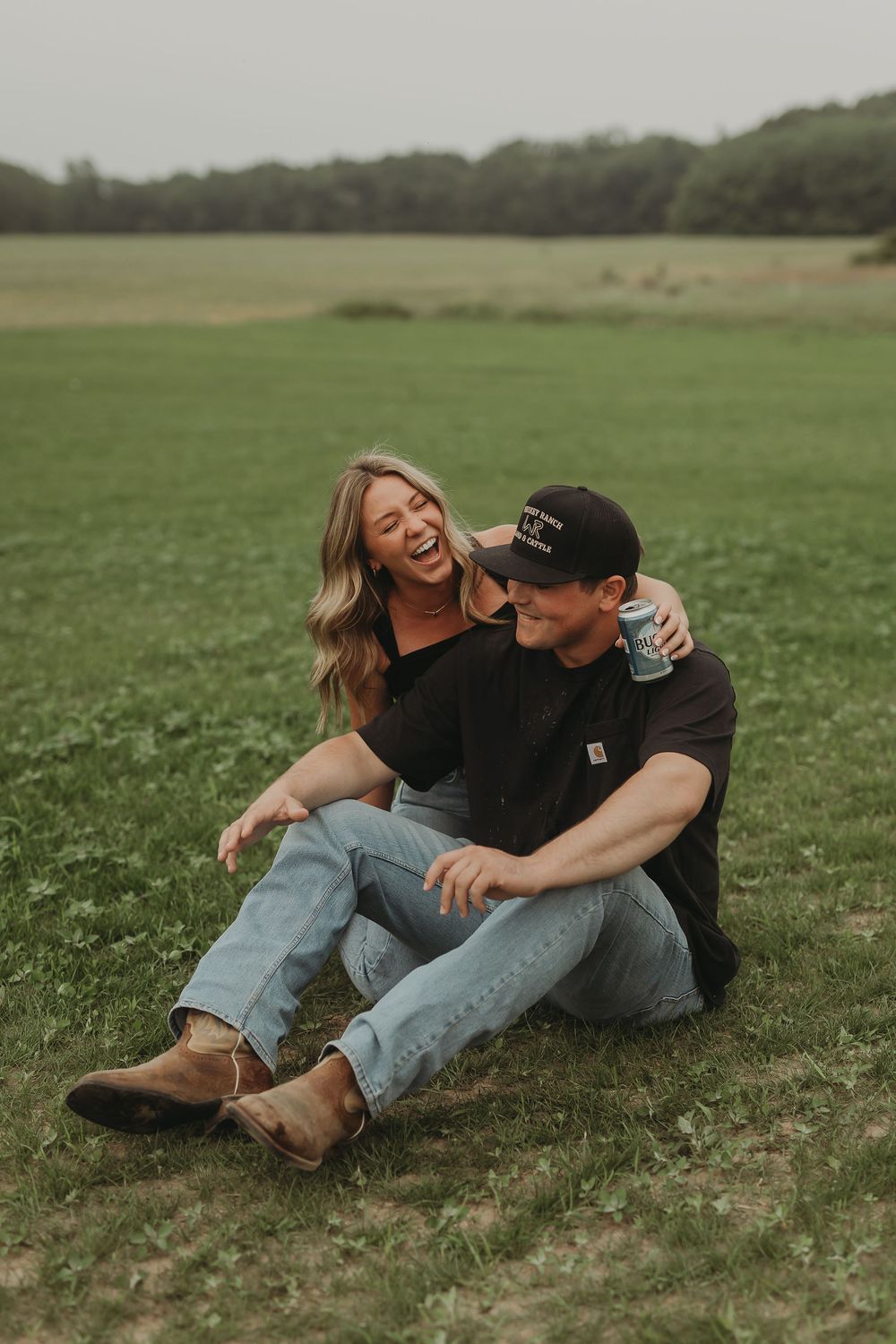 A couple sitting and laughing together on green grass in a field during an overcast day.