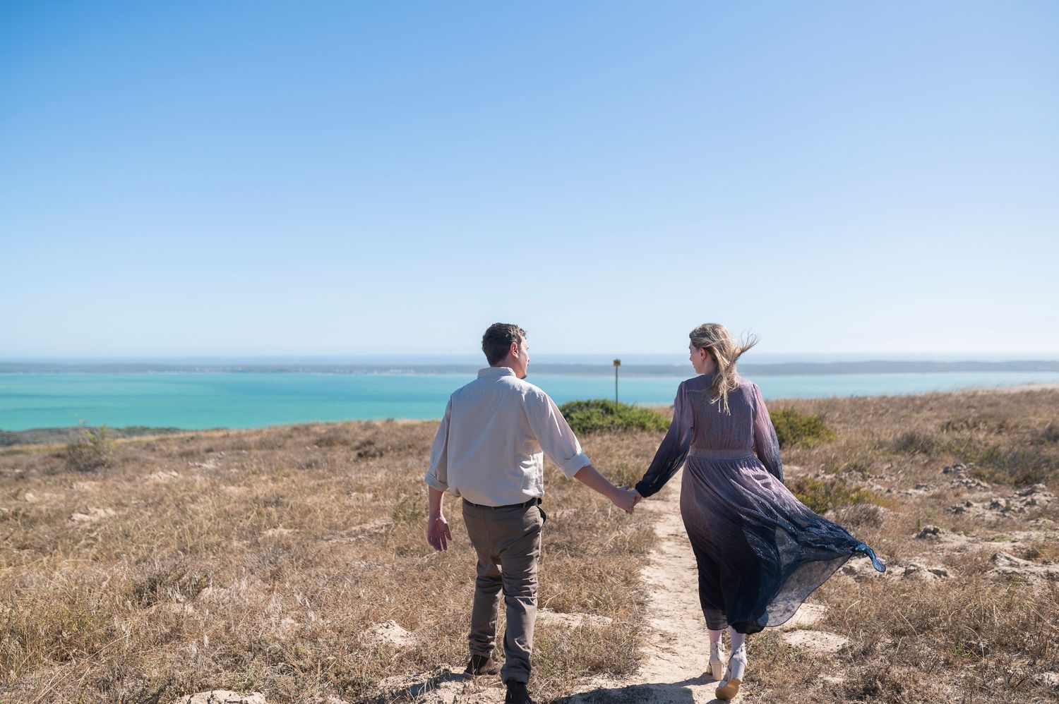 A couple holding hands walks along a coastal cliff overlooking turquoise ocean waters on a sunny day.