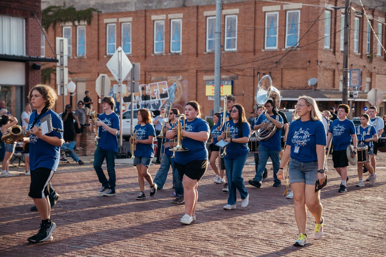 A marching band wearing blue uniforms performs on brick pavement during a parade or street performance.