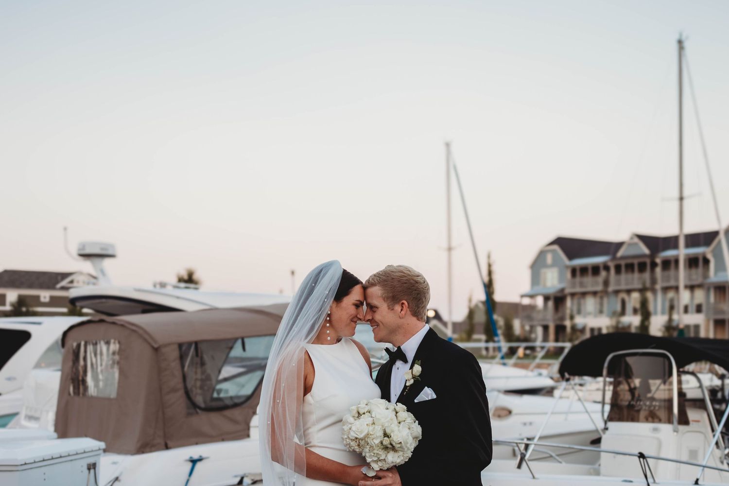 A romantic nautical wedding portrait of a couple embracing on a dock with boats and marina buildings in the background.