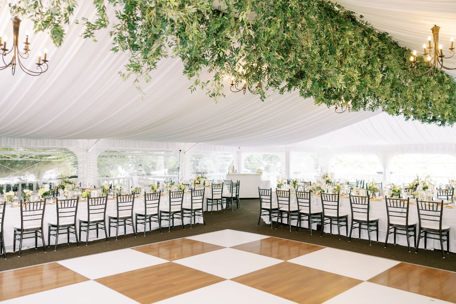 White wedding tent interior with elegant table settings, chiavari chairs, and lush greenery draped from the ceiling.