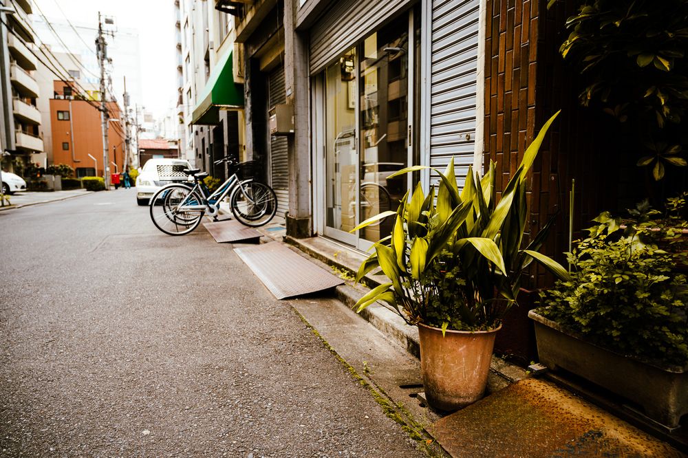 Narrow alley in Japan with potted plants and bicycles lined against traditional buildings on a rainy day.