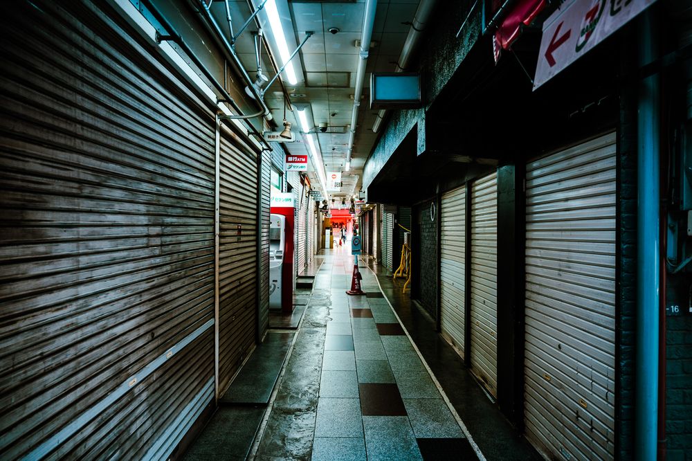 Empty shopping arcade corridor with metal shutters and fluorescent lighting creates an eerie atmosphere.