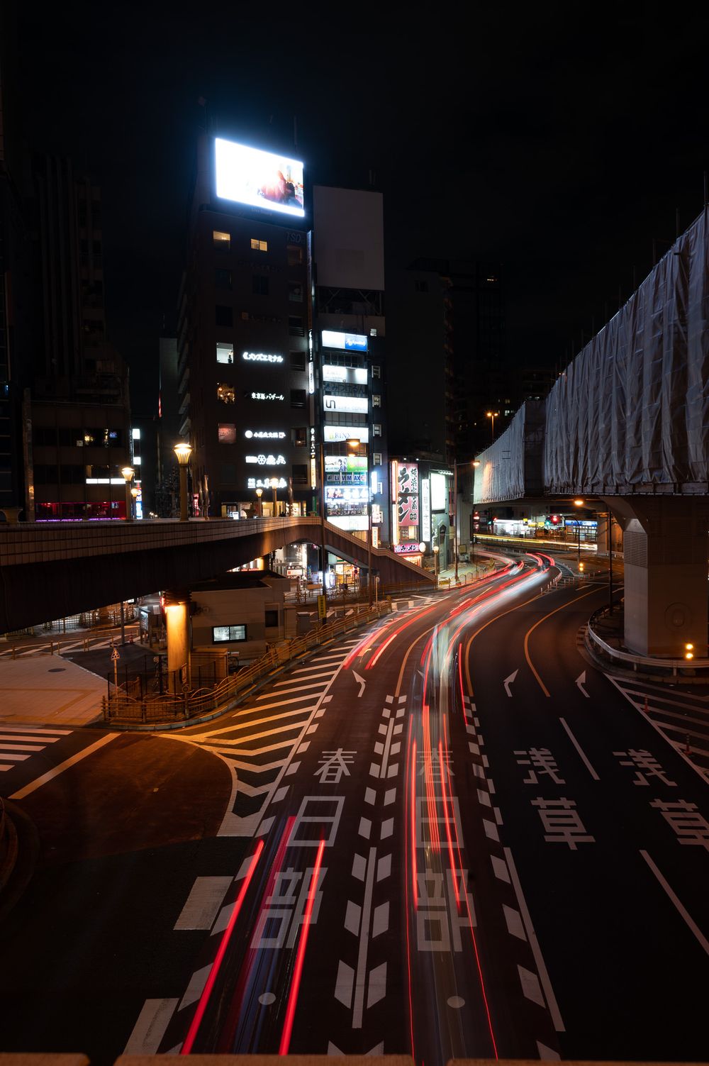 Long exposure nighttime photograph showing light trails from cars on a curving city street with illuminated buildings in Japan.