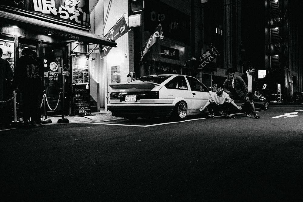 Black and white street photograph of buildings and cars in an urban alley at night with dramatic shadows.