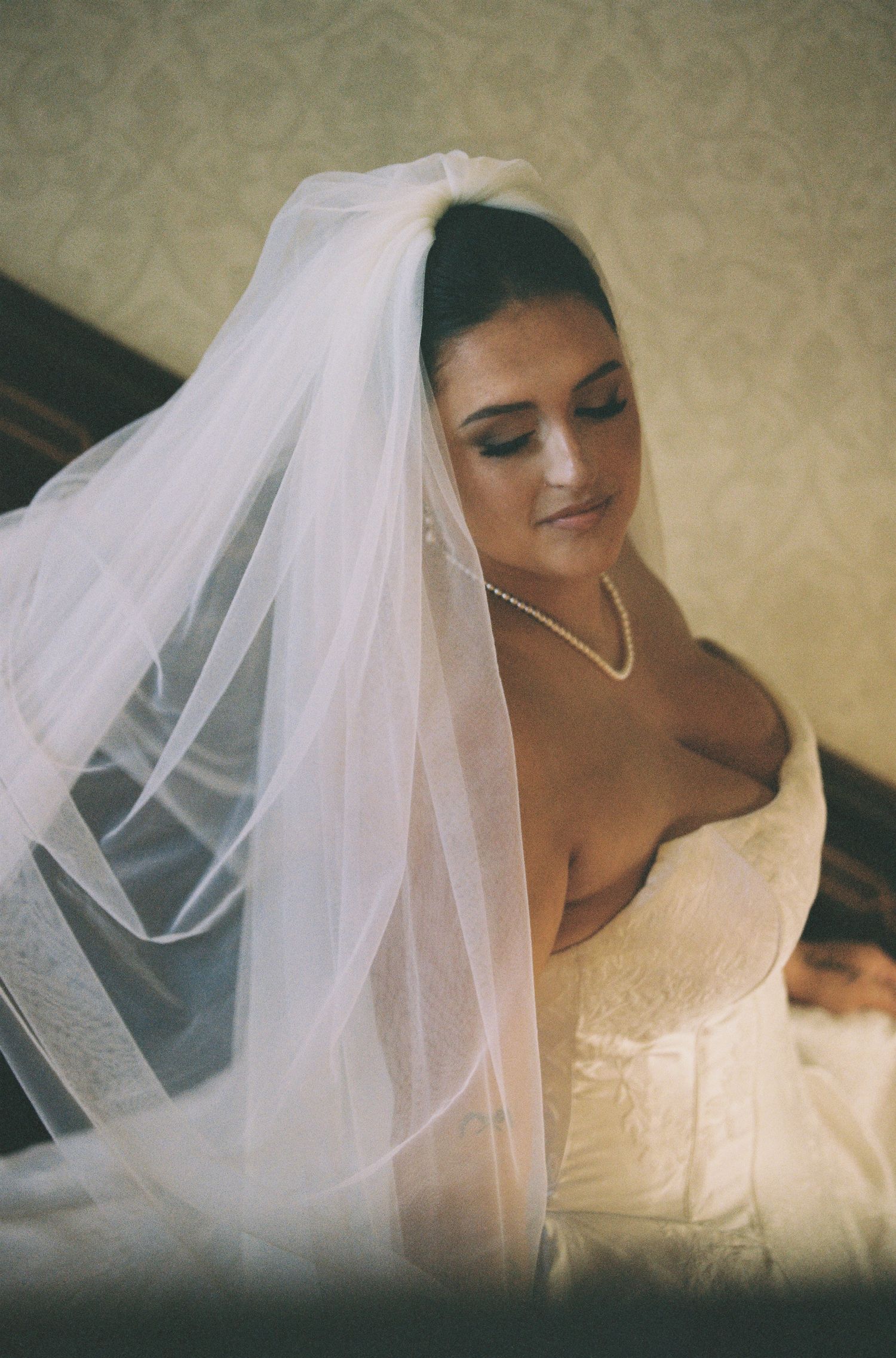 Elegant bride in a white lace gown and pearl necklace, draped in a flowing veil, gazing downward softly.