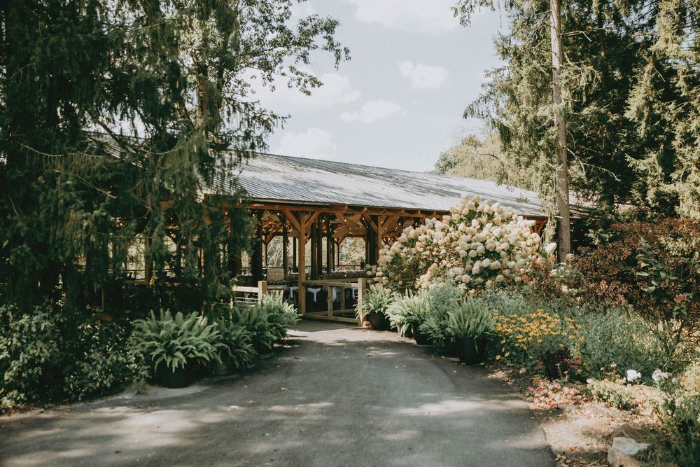A rustic wooden pavilion surrounded by lush gardens and trees along a paved path on a sunny day.