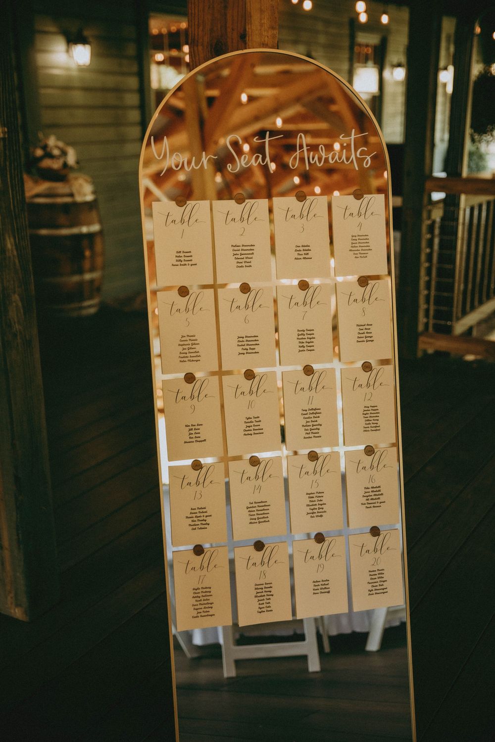 A gold-framed mirror wedding seating chart displays handwritten table assignments on kraft paper cards in an elegant setup.