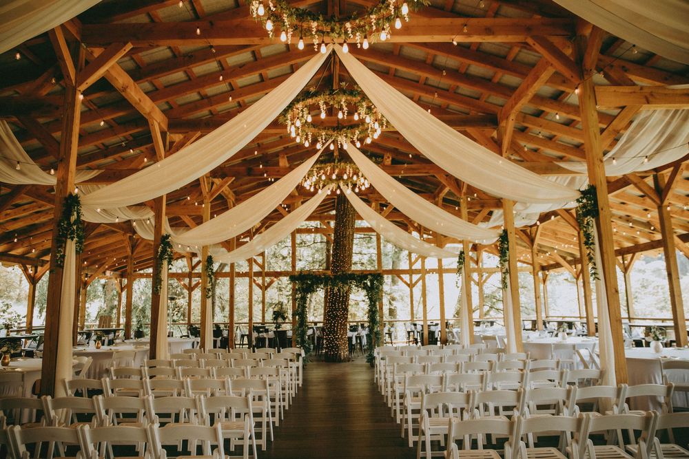 Rustic wooden beams and elegant chandeliers decorate an indoor wedding venue set up with white chairs for a ceremony.