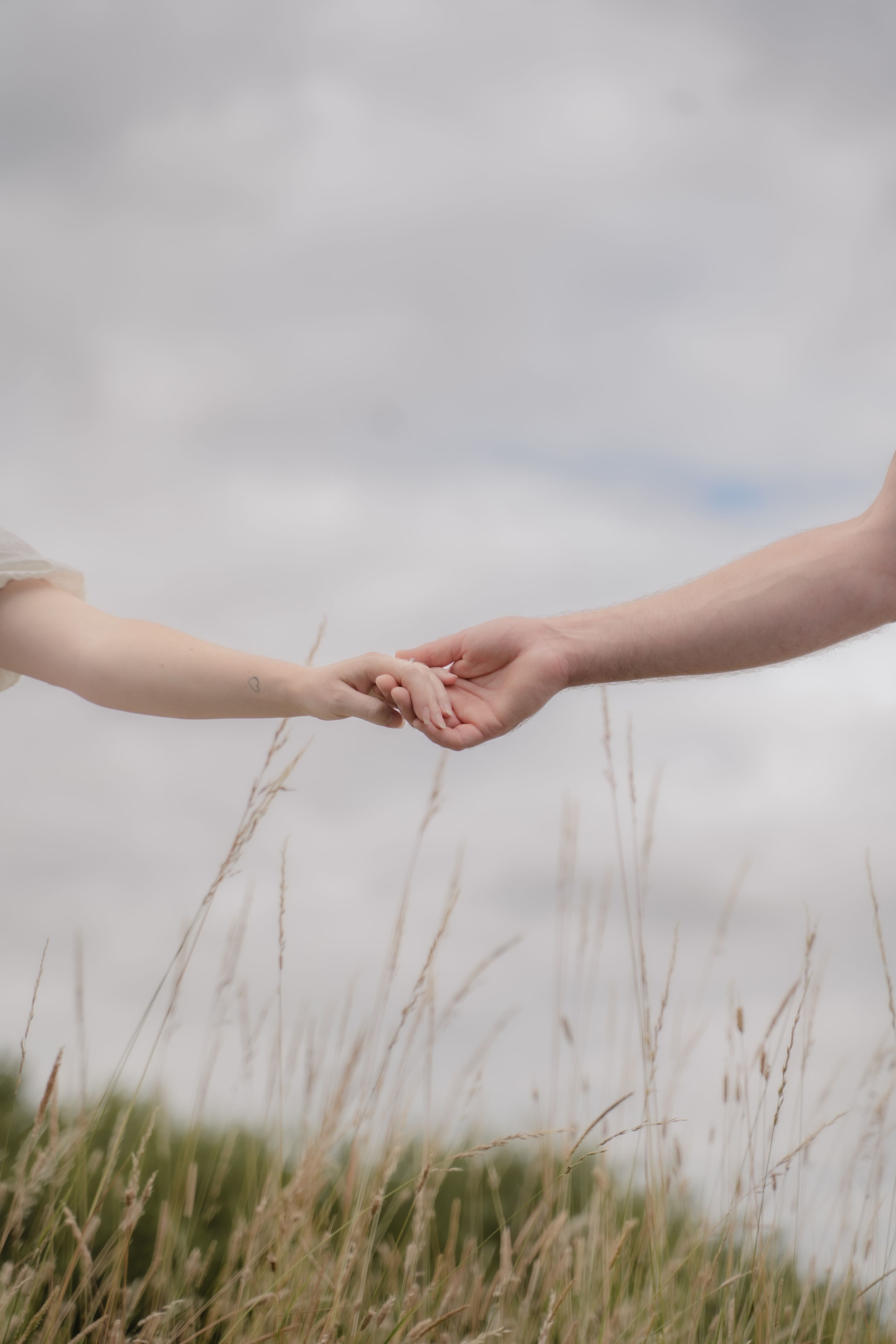 Two hands reaching to touch each other in a tender moment above tall grass against a cloudy sky.