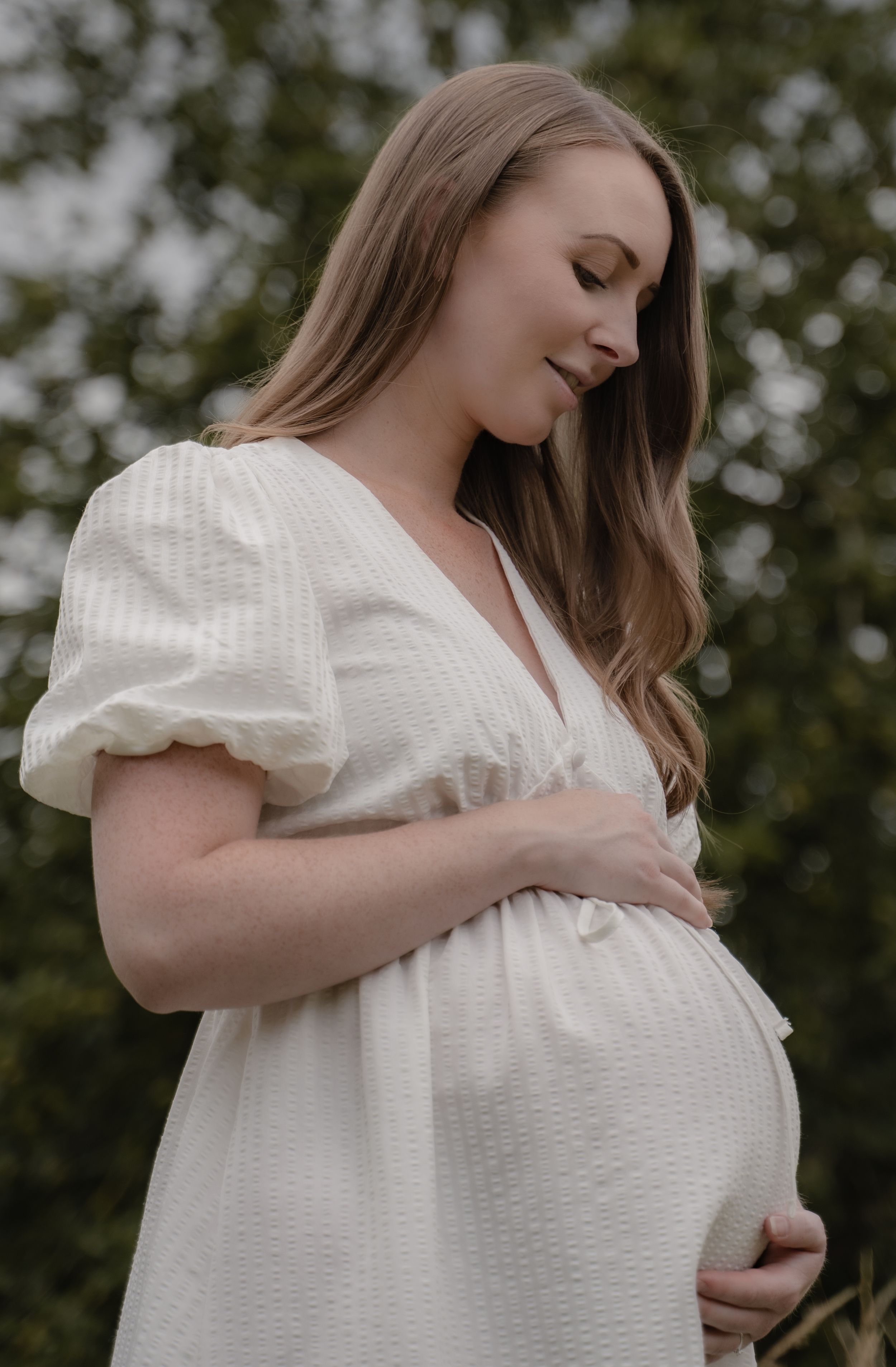 A pregnant woman in a white dress with puff sleeves cradles her belly while standing outdoors in a peaceful moment.