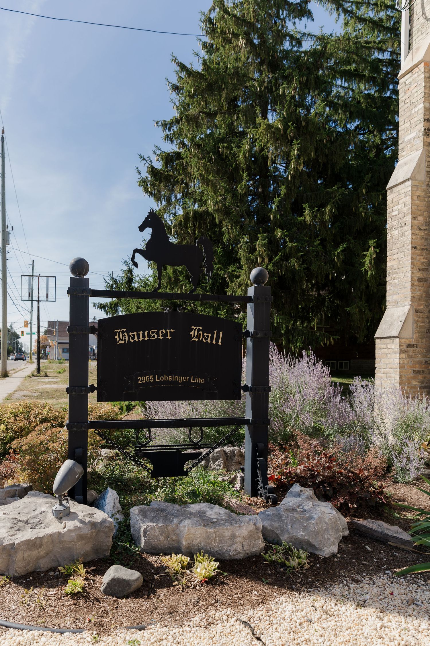 Outdoor entrance sign for Hauser Hall in Kitchener, Ontario with landscaped rock garden and evergreen trees.
