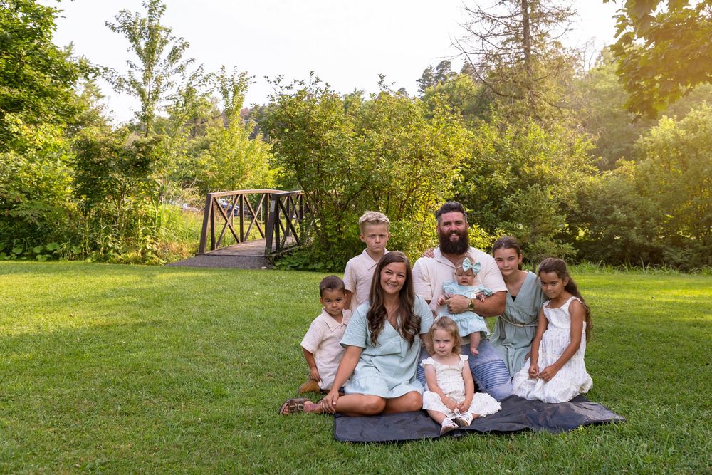 A family of 8 sitting on a blanket smiling at the camera at Orono Park with a bridge behind them in the background