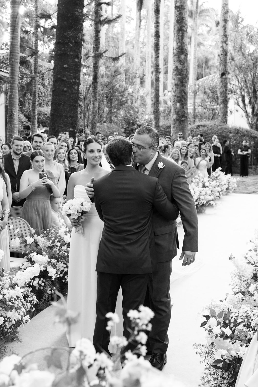Black and white photo of a wedding ceremony with flower-lined aisle and gathered guests.