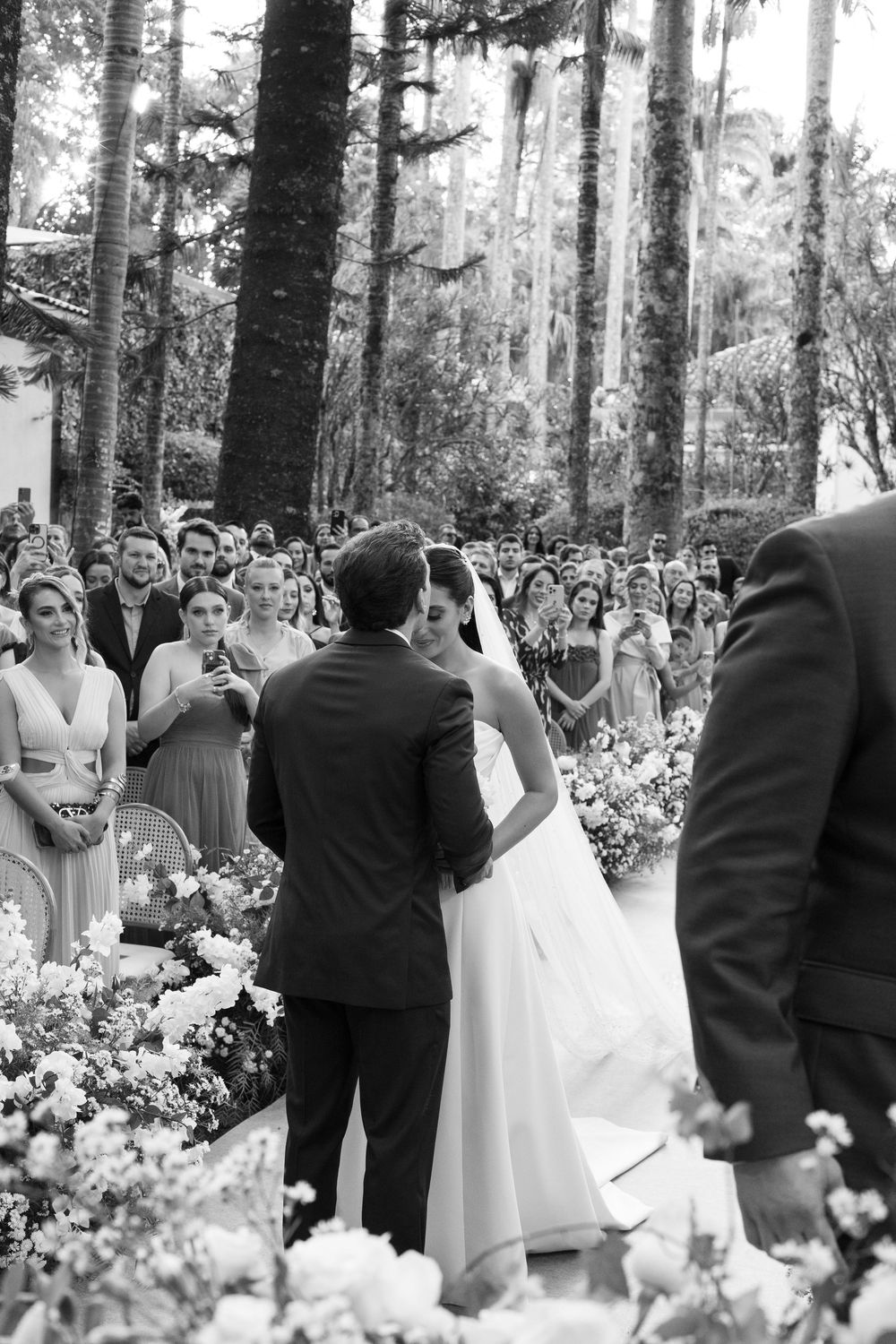 A black and white photo of a wedding ceremony surrounded by tall trees and flowers.