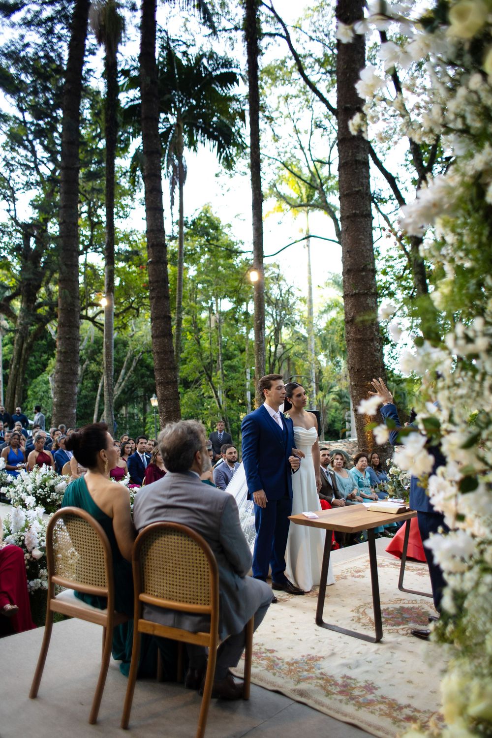 Outdoor woodland wedding ceremony setup with rustic wooden chairs and white floral decor among tall pine trees.