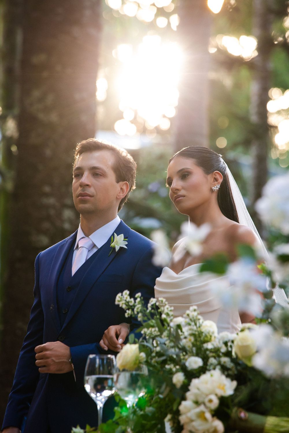 Romantic wedding couple shares intimate moments under sunlit trees with glowing bokeh and white floral decorations.