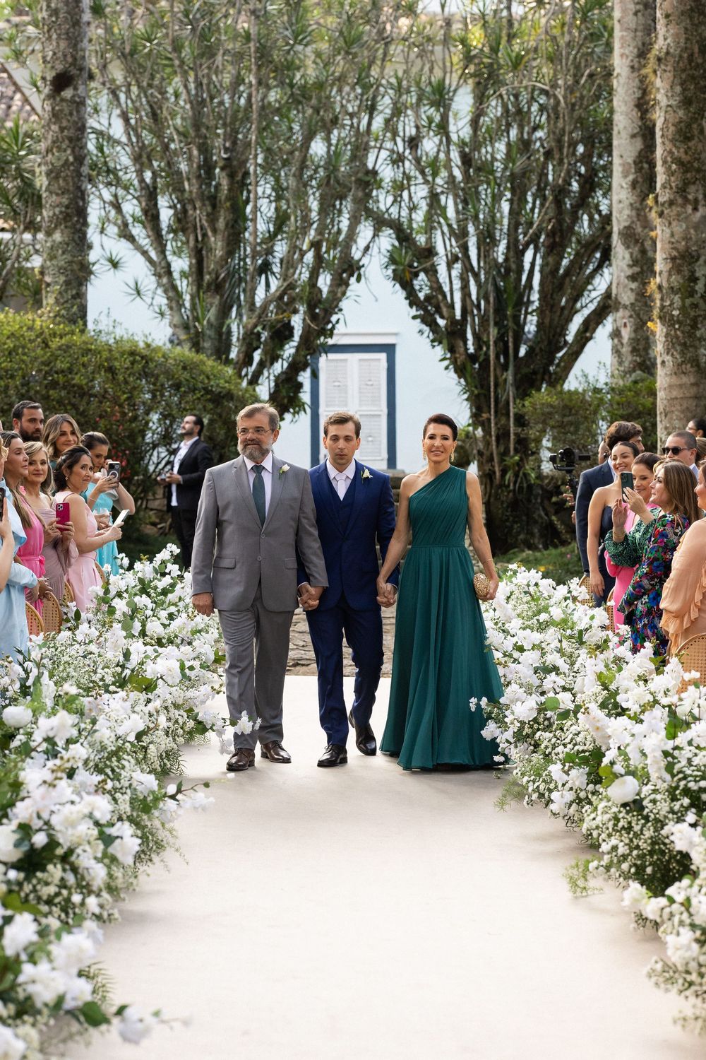 Wedding guests line a path adorned with white flowers and tall trees during an outdoor ceremony.