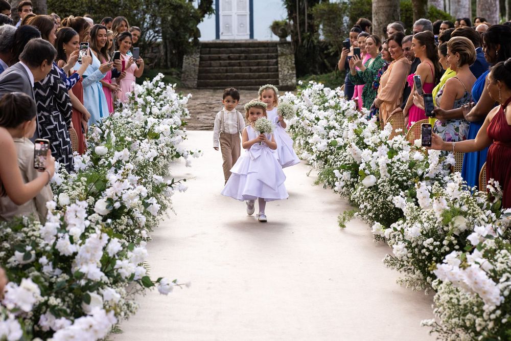 Young flower girls walk down an outdoor aisle lined with white floral arrangements as wedding guests look on from both sides.