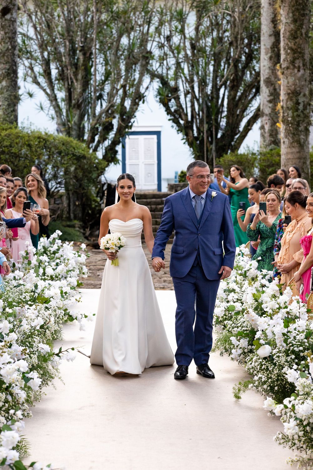 Wedding processional on stone pathway lined with white flowers as guests watch from both sides.