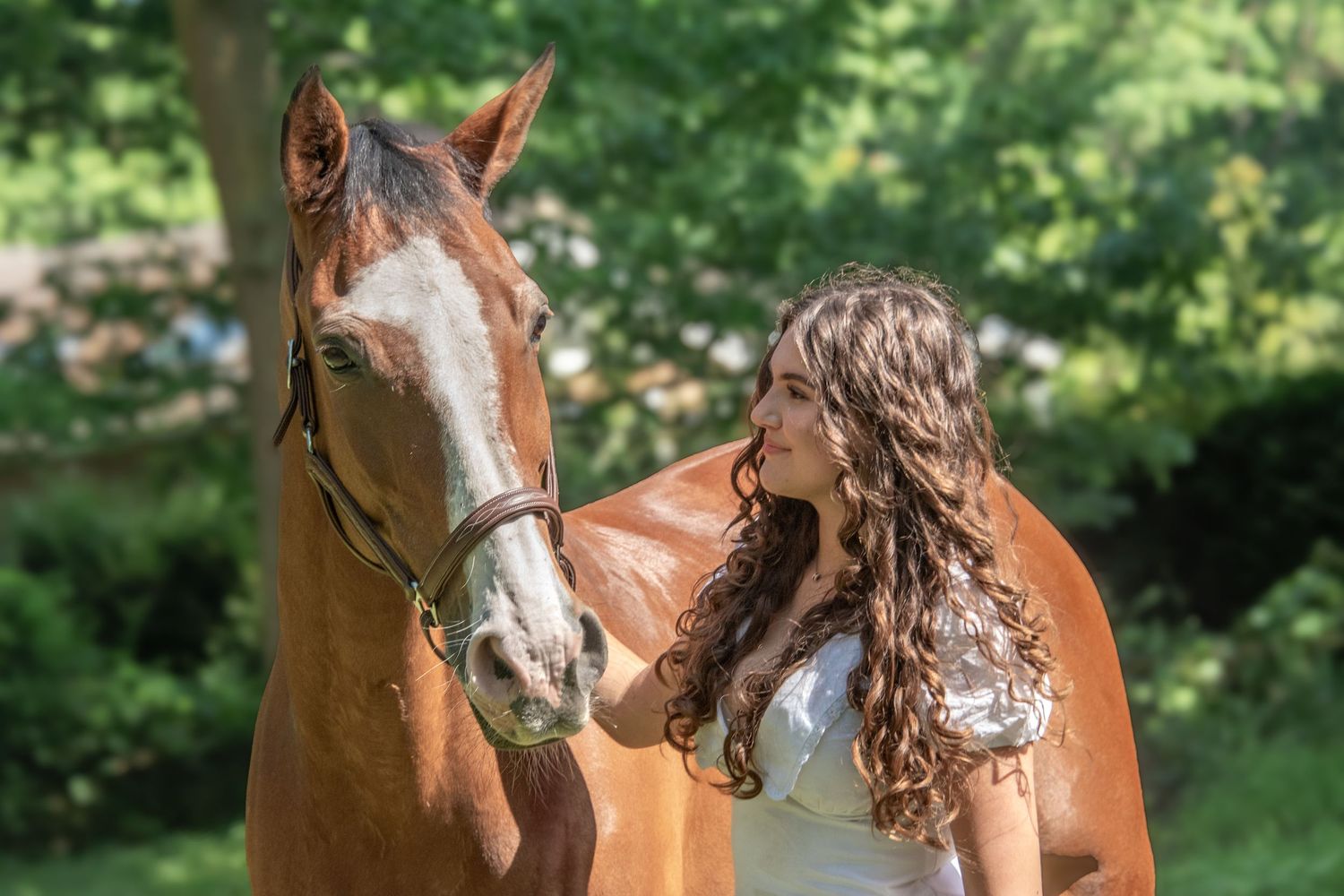 Young woman with curly hair in white dress standing beside a chestnut horse outdoors.